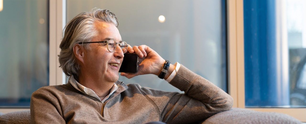 Man in a beige sweater is sitting on the couch and has a phone in his left hand